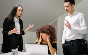 Mulher sentada em frente ao computador com sinais de estresse devido a pressão no trabalho rodeada por pessoas.